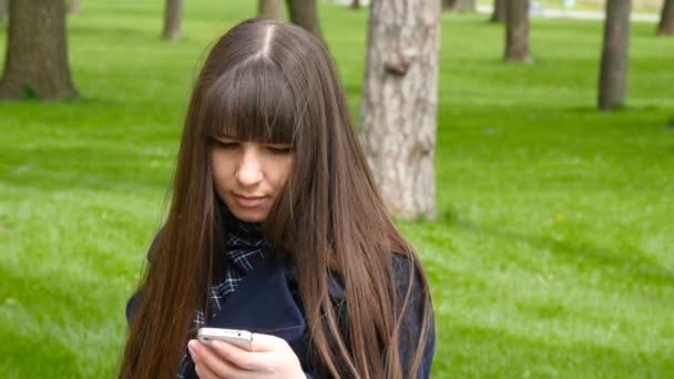 美丽的女人用细胞智能手机在户外公园-详细信息。年轻有魅力的快乐的女孩在城市公园的放松和使用手机。她看起来很快乐也很满足 — 图库视频影像