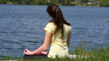 Genç kadın virajlı baş to-and-fro egzersizleri yapıyor. Çimenlerin üzerinde oturan kız ve lotus pozisyonu sırasında yoga egzersizleri yapıyor nehir yanında meditasyon