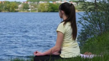 Meditasyon ve smartphone içinde lotus pozisyonu kulaklık müzik dinleme genç kadın. Çimenlerin üzerinde oturan ve nehrin yanında Sahilde meditasyon rahatlatıcı kız