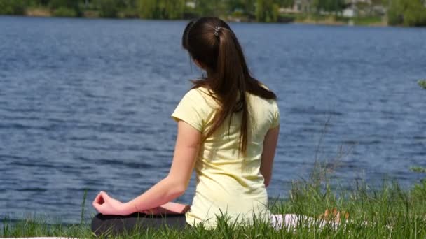 Mladá žena dělá cvičení ohýbá hlavou fro. Dívka sedící v trávě a meditují vedle řeky dělat cvičení jógy v lotosové pozici — Stock video