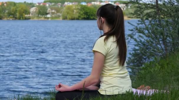 Jovem mulher meditando e ouvindo música no smartphone em fones de ouvido na posição de lótus. Menina sentada na grama e relaxante meditando na praia ao lado do rio — Vídeo de Stock