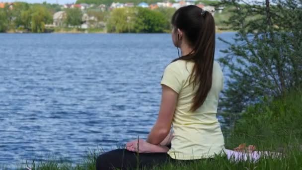 Mujer joven meditando y escuchando música en un smartphone con auriculares en posición de loto. Chica sentada en la hierba y relajada meditando en la playa junto al río — Vídeo de stock