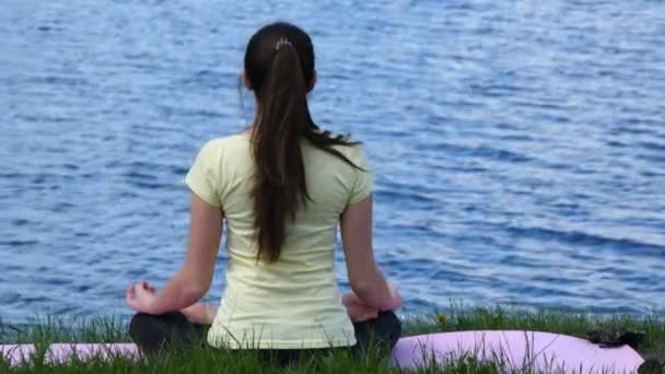 Jovem mulher relaxante meditando na praia. Menina sentada na grama e meditar ao lado do rio fazendo exercícios de ioga durante a posição de lótus — Vídeo de Stock