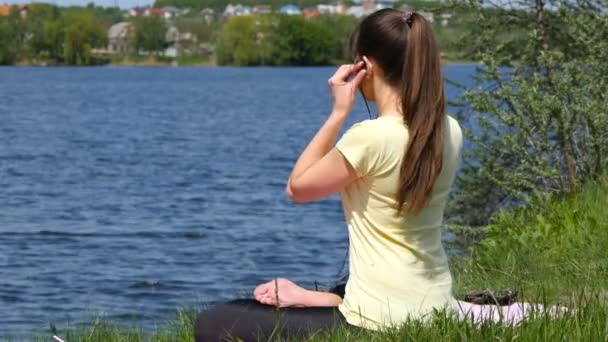 若い女性は彼女の耳にイヤホンを入れて、蓮華座で瞑想します。女の子の芝生に座って、川沿いにビーチで瞑想リラックス — ストック動画