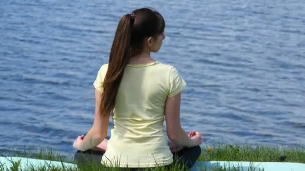 Mujer joven hace ejercicios dobla la cabeza de un lado a otro. Chica sentada en la hierba y meditar junto al río haciendo ejercicios de yoga durante la posición de loto — Vídeo de stock