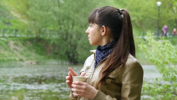 Hermosa joven en el parque bebiendo una taza de café para llevar de una taza desechable. Retrato de mujer atractiva cuidadosamente mira hacia fuera sobre el río y la celebración de la taza de té para calentar — Vídeos de Stock