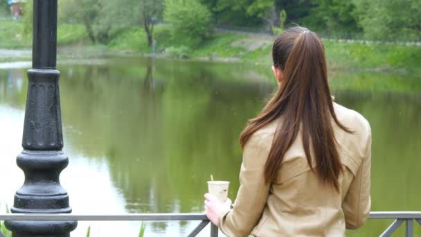 使い捨てのカップからコーヒーのテイクアウトのカップを飲む少女の背面します。女性は思慮深く川と茶はウォーム アップのホールディング カップを眺めること — ストック動画