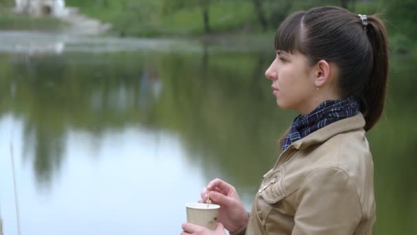 Kaunis nuori tyttö seisoo rantakadulla joen ja juominen kuppi takeaway kahvia kertakäyttöinen kuppi — kuvapankkivideo