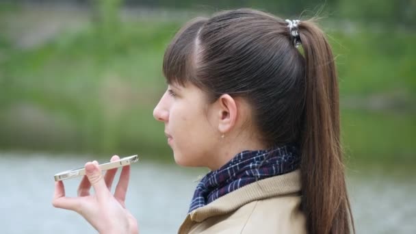 Una mujer joven se para en el terraplén del río y habla por teléfono celular. Chica hablando en el teléfono inteligente al aire libre en primavera. Primer plano — Vídeo de stock