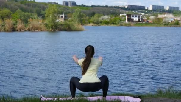 Dziewczynka fitness ćwiczenia na świeżym powietrzu nad rzeką kucać ćwiczenie w niesamowity krajobraz natura. Dopasowanie młoda kobieta robi kuca na matę na plaży. Widok z tyłu — Wideo stockowe