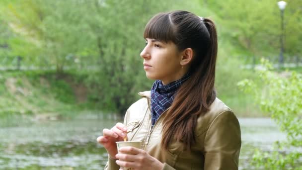 Hermosa joven en el parque bebiendo una taza de café para llevar de una taza desechable. Retrato de mujer atractiva cuidadosamente mira hacia fuera sobre el río y la celebración de la taza de té para calentar — Vídeos de Stock