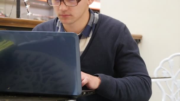 Молодой человек в очках сидит за столом в торговом центре и работает на ноутбуке — стоковое видео