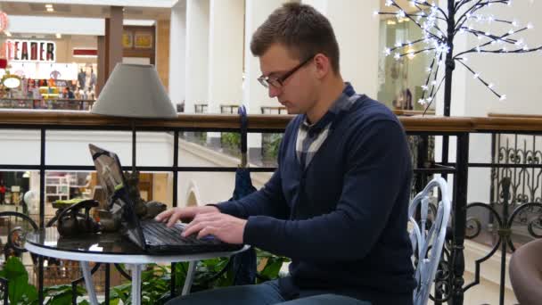 Hombre joven en gafas están sentados en la mesa en el centro comercial y trabajando en un ordenador portátil — Vídeos de Stock