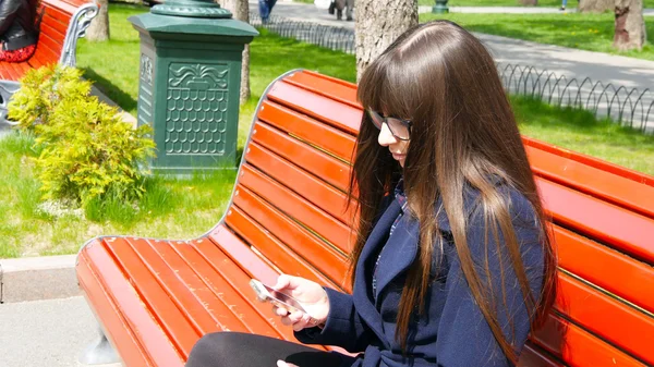 Mujer joven en gafas utiliza teléfono inteligente celular en un parque de la ciudad. Chica sentada en un banco rojo al aire libre en primavera y utiliza un teléfono móvil. Primer plano —  Fotos de Stock
