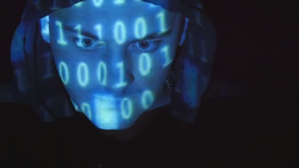 Hooded hacker working on a computer, binary code projecting on his face. Source code projected over an angry hostile mans face, black background. — Stock Video