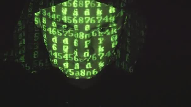 面对男性黑客在罩在计算机上工作，而绿色代码字符反映在他的脸在黑暗的办公室里。源代码上投射下来生气的敌对的男人的脸，黑色背景. — 图库视频影像