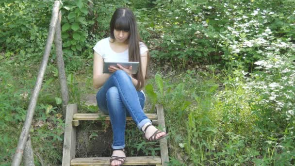 Hermosa joven sentada en las escaleras de madera en el bosque y el uso de tableta pc. Chica feliz navegar por Internet, el uso de la aplicación y la comunicación al aire libre. Relax y tecnología . — Vídeos de Stock