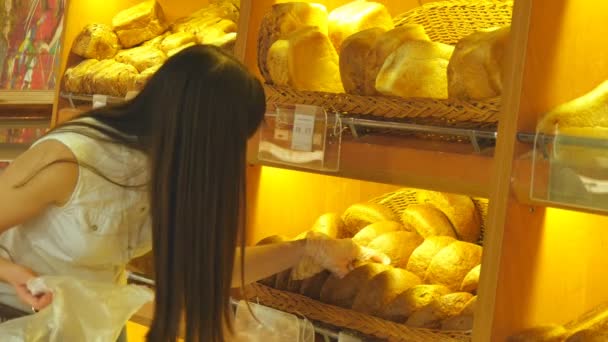Женщина выбирает и кладет свежий хлеб в пакет в супермаркете. Молодая девушка берет буханку хлеба с полки, нюхает его и кладет в корзину. Покупки в продуктовом магазине — стоковое видео