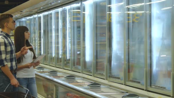 Pareja joven está parada junto al congelador en la tienda de comestibles y recogiendo paquetes con alimentos congelados. El hombre cogiendo el producto de la nevera. Mujer usando la tableta PC para comprobar la lista de compras — Vídeos de Stock