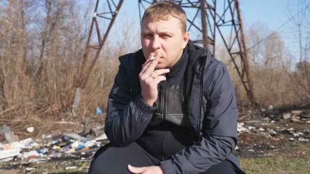 Adult serious man sits squatting and smoking cigarette outdoor. Male person looking into camera and throwing out a cigarette butt. Concept of unhealthy habit and addiction. Close up Slow motion — Video Stock