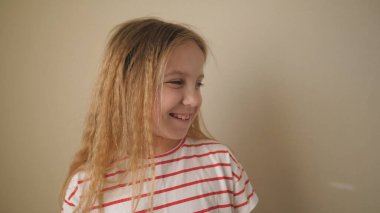 Küçük gülümseyen kız çocuğu yan tarafa bakıyor ve içeride biriyle gülüyor. Yüzünde tatlı bir çocuğun olumlu duyguları var. Duvarın arkasında mutlu küçük sarışın kız. Yavaşla..