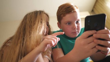 Küçük çocuklar odasındaki kanepede oturmuş akıllı telefondan ilginç videolar izliyorlar. Kız ve erkek kardeş evde cep telefonuyla film izliyorlar. Küçük oğlan ve kız birlikte vakit geçiriyorlar. Kapat..