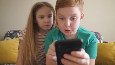 Küçük çocuklar odasındaki kanepede oturmuş akıllı telefondan ilginç videolar izliyorlar. Kız ve erkek kardeş evde cep telefonuyla film izliyorlar. Küçük oğlan ve kız birlikte vakit geçiriyorlar. Kapat..