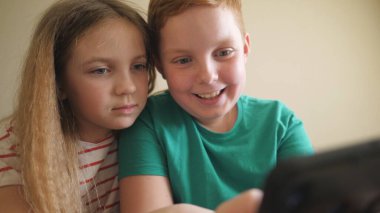 Neşeli küçük oğlan ve kız odasındaki kanepede oturan akıllı telefondan komik bir video izliyorlar. Küçük gülümseyen erkek ve kız kardeş evde cep telefonuyla oyun oynuyor. Çocuklar birlikte vakit geçiriyor..