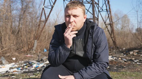 Adult serious man sits squatting and smoking cigarette outdoor. Male person looking into camera and throwing out a cigarette butt. Concept of unhealthy habit and addiction. Close up Slow motion.