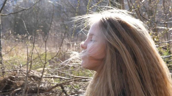 ケアフリーの女の子は頭を上げて 長いブロンドの髪を空中で振っています 早春の森のぼやけた背景にカメラを向ける美しい女性の子供の肖像画 スローモーション — ストック写真