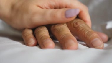 Torun, hasta yetişkin bir büyükannenin yatakta yatan kırışık koluna nazikçe dokunur. Kız klinikte yaşlı büyükannesinin elini rahatlatıyor. Kızı annesine destek oluyor.