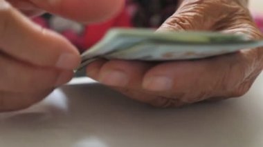 Yaşlı bir kadın nakit parayı elinde tutuyor ve masanın üzerinde döviz sayıyor. Yaşlı büyükannenin ellerini kapat ve 100 dolarlık banknotları masaya koy. Para konsepti. Düşük görünüm Yavaş çekim
