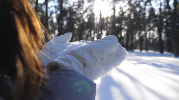 白い手袋の中の認識できない女性は彼女の手のひらから雪を吹いている。冬の森の中に立つ若いブルネットの女の子と晴れた日に雪と遊ぶ。背景がぼやけている。スローモーション閉じる — ストック動画