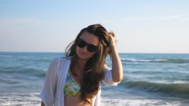 Güneş gözlüklü çekici bayan deniz manzaralı arka planda gülümsüyor ve okyanus kıyısında geziniyor. Keyfi yerinde bir kadının portresi özgürlüğün tadını çıkarıyor ve sahilde eğleniyor. Tatil köyünde eğlenen tatlı kız.