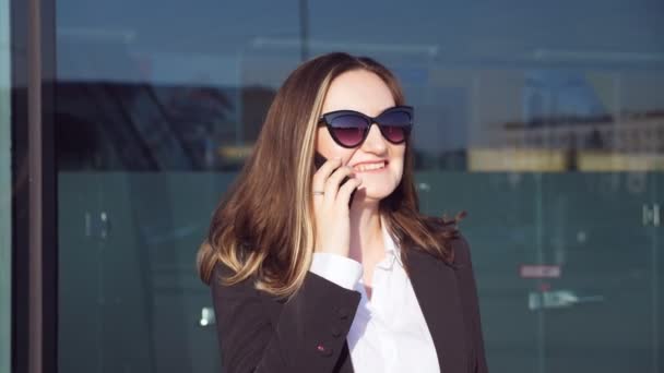 Egy fiatal, mosolygós üzletasszony portréja, aki telefonon beszél. Boldog gyönyörű nő beszél okostelefonon közelében modern üzleti központ. Az üzletasszony jó hangulatban van. Lassú mozgás Bezárás — Stock videók