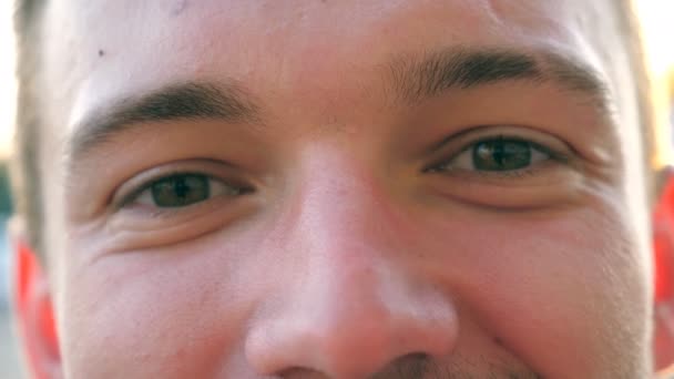 Green eyes of young confident guy blinking and looking into camera at city street. Portrait of happy handsome man smiling in urban environment at sunset. Sunlight at background. Close up — Stock Video