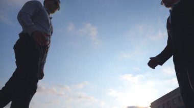 İki başarılı iş adamı şehirde buluşup birbirlerini selamlıyorlar. Genç erkek meslektaşlar dışarıda el sıkışıyor. İş ortaklarının el sıkışması. Arka planda mavi gökyüzü. Yavaş çekim Düşük açı görünümü