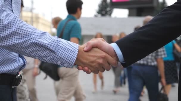 Два бизнесмена приветствуют друг друга в городской среде. Молодые коллеги-мужчины пожимают друг другу руки с размытым городским прошлым. Рукопожатие деловых партнеров на открытом воздухе. Закрыть медленное движение — стоковое видео