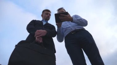 Genç iş adamı, şehirdeki meslektaşına tablet ekranında bir sunum gösteriyor. Başarılı girişimciler arka planda mavi gökyüzü olan dijital tablet üzerinde çalışıyorlar. Düşük açı görünümü Yavaş çekim