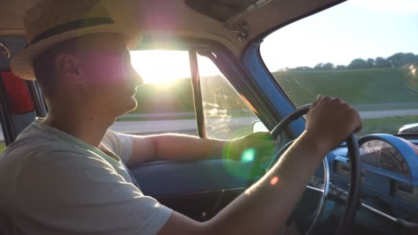 戴帽子的年轻人手牵着方向盘，开着一辆带有太阳耀斑的复古汽车。在乡村乘坐老式汽车旅行的白人。侧视慢动作闭合 — 图库视频影像