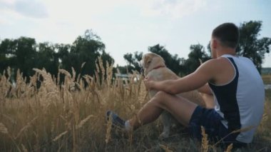 Genç adam çimde oturur ve labradorunu ya da golden retriever 'ını okşar. Adam doğada evcil hayvanıyla vakit geçiriyor. Köpeği olan beyaz bir erkek, dinlenmenin tadını çıkarıyor. Yan görünüm Yavaş çekim