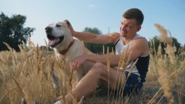 Genç adam çimde oturur ve labradorunu ya da golden retriever 'ını okşar. Adam doğada evcil hayvanıyla vakit geçiriyor. Köpeği olan beyaz bir erkek, dinlenmenin tadını çıkarıyor. Yavaş çekim Dolly.