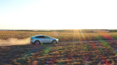 Elektrikli aracın hava görüntüsü off-road yolunda ilerlerken ardında toz izi bırakıyor. Kırsal yol boyunca yeni bir araba sürüşü. Ekoloji dostu araç elektrik yüküne biniyor. Arka planda güneşli manzara.