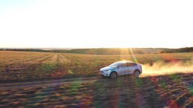 Elektrikli aracın hava görüntüsü off-road yolunda ilerlerken ardında toz izi bırakıyor. Kırsal yol boyunca giden elektrik yükündeki ekoloji dostu otomobil. Arka planda manzaralı günbatımı manzarası. Yan görünüm.