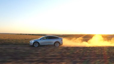 Tozlu yoldan hızla geçen elektrikli arabanın havadan görüntüsü. Ekoloji dostu otomobil, arka planda günbatımı manzaralı kırsal yolda gidiyor. Modern teknoloji kavramı.
