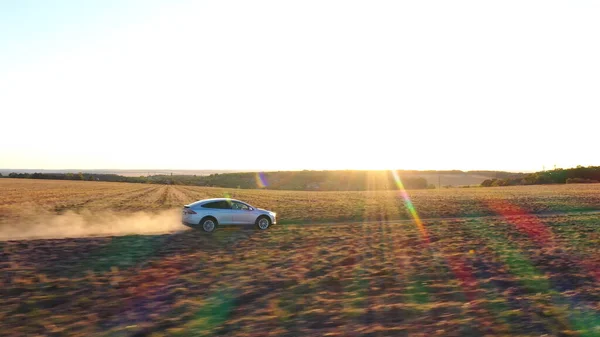 Aerial shot of electrical car moving on off-road route leaving dust trail behind. Ecology friendly auto on electric charge driving along rural road. Scenic sunset landscape at background. Side view.