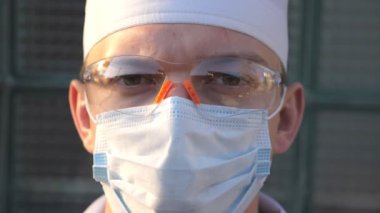 Tıbbi maske takan beyaz erkek doktorun portresi kameraya bakıyor. Virüsten koruyucu maske takan bir tıp çalışanı. Koronavirüs salgınından sağIık ve güvenlik kavramı. COVID-19