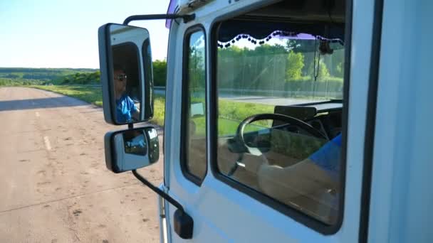 Camionero en gafas de sol que reflejan en el espejo lateral del coche en movimiento. Hombre conduciendo camión a caballo a destino a través de carretera rural. Concepto de transporte de carga. Hermoso fondo. Lento mo Primer plano — Vídeo de stock