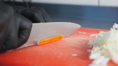 Eldivenli aşçının erkek elleri mutfak restoranında bıçakla plastik doğrama tahtasındaki yengeç çubuğunu dilimliyor. Sağlıklı bir sebze salatası hazırlama süreci. Yemek pişirme kavramı. Yavaş çekim