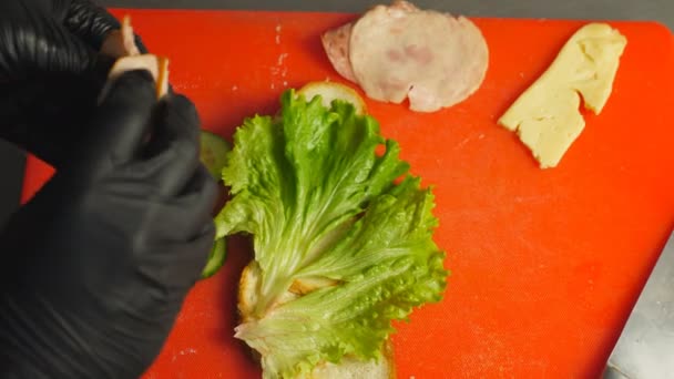 Chef legt Stücke von Schinken, Salami und Käse auf Brot mit Salatblatt in der Küche. Männliche Hände in Handschuhen kochen in der Küche ein leckeres Sandwich. Prozess der Zubereitung schmackhafter Snacks. Ansicht von oben — Stockvideo